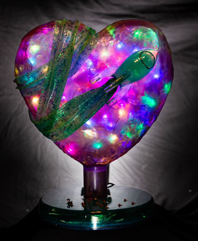 A large purple heart cast from epoxy resin. The heart is held up by a blue pedestal with little ducks on the pond. A turquoise Fairy Dust Rocket flies past the heart, pulling a glittery bandage over it. 
        Photo (c) c3awareness | TRim