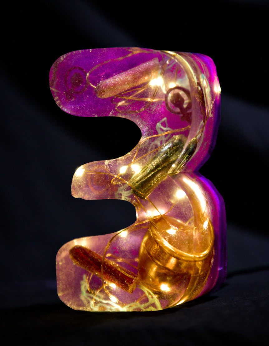 A cast of the letter 3 made of epoxy resin. Inside, a string of lights glows, and tampons covered in glitter nail polish and a golden menstrual cup float around. Next to it are feminist symbols of a purple fist in a circle. The letter is coloured pink purple. Photo (c) c3awareness | TRim