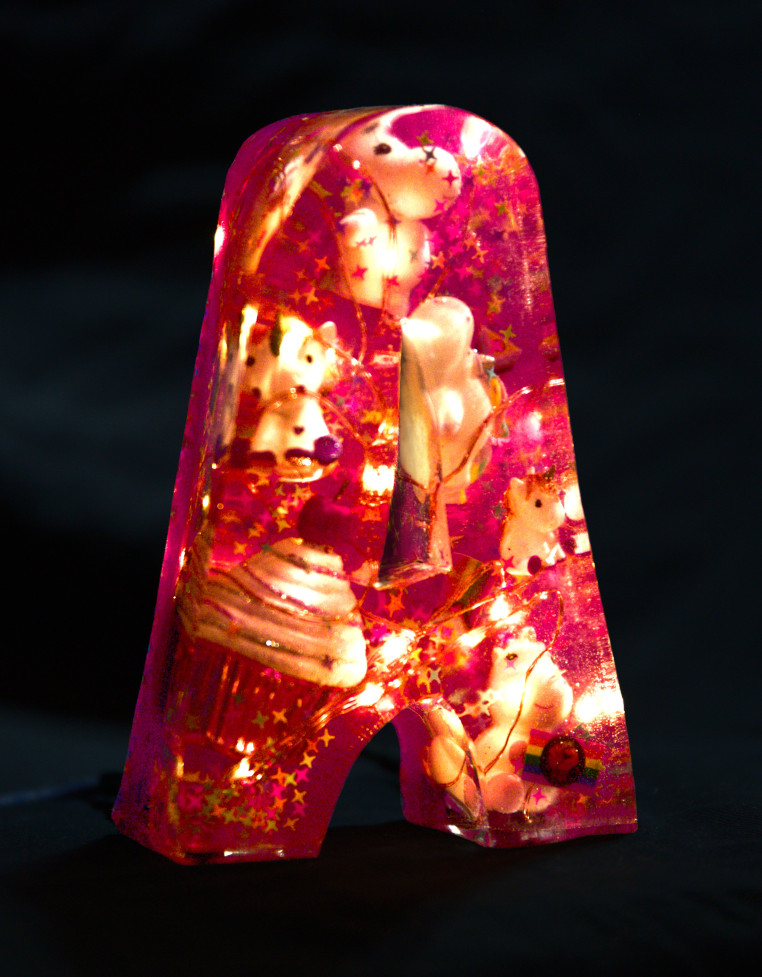 Letter 'A' moulded from epoxy resin. Inside, a string of lights glows and unicorns, rainbow flags, a cupcake with a cherry and decorative stars float around. The letter is coloured pink. Photo (c) c3awareness | TRim