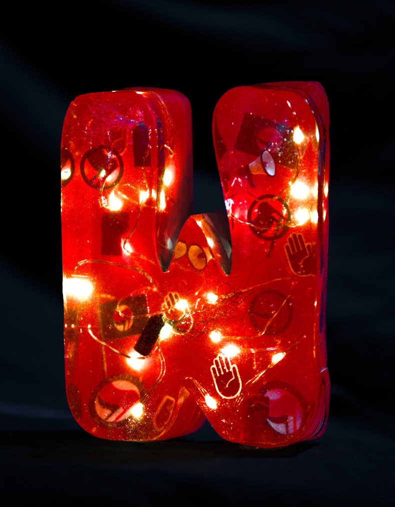 Letter 'W' moulded from epoxy resin. Inside, a string of fairy lights glows and anti-fascist flags, megaphones, spray cans in red and black, and red hands signing 'stop' float around. The letter is coloured red. Photo (c) c3awareness | TRim