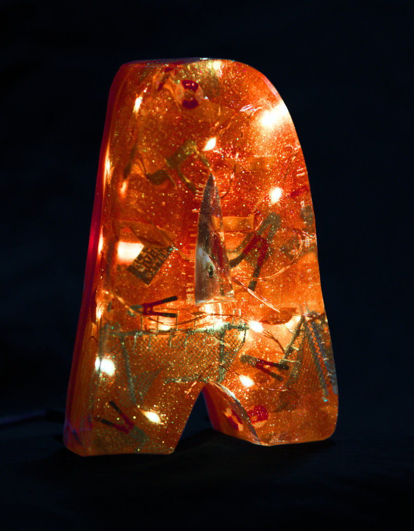 An 'A' made of epoxy resin. Inside, some fairy lights glow and small folded paper boats, lifebuoys, wire representing cut border fences, small bolt cutters, flags with ‘Black lives matter’ and the Hebrew word ‘Chai’ as a Jewish symbol for life float inside. The letter is coloured orange. Photo (c) c3awareness | TRim