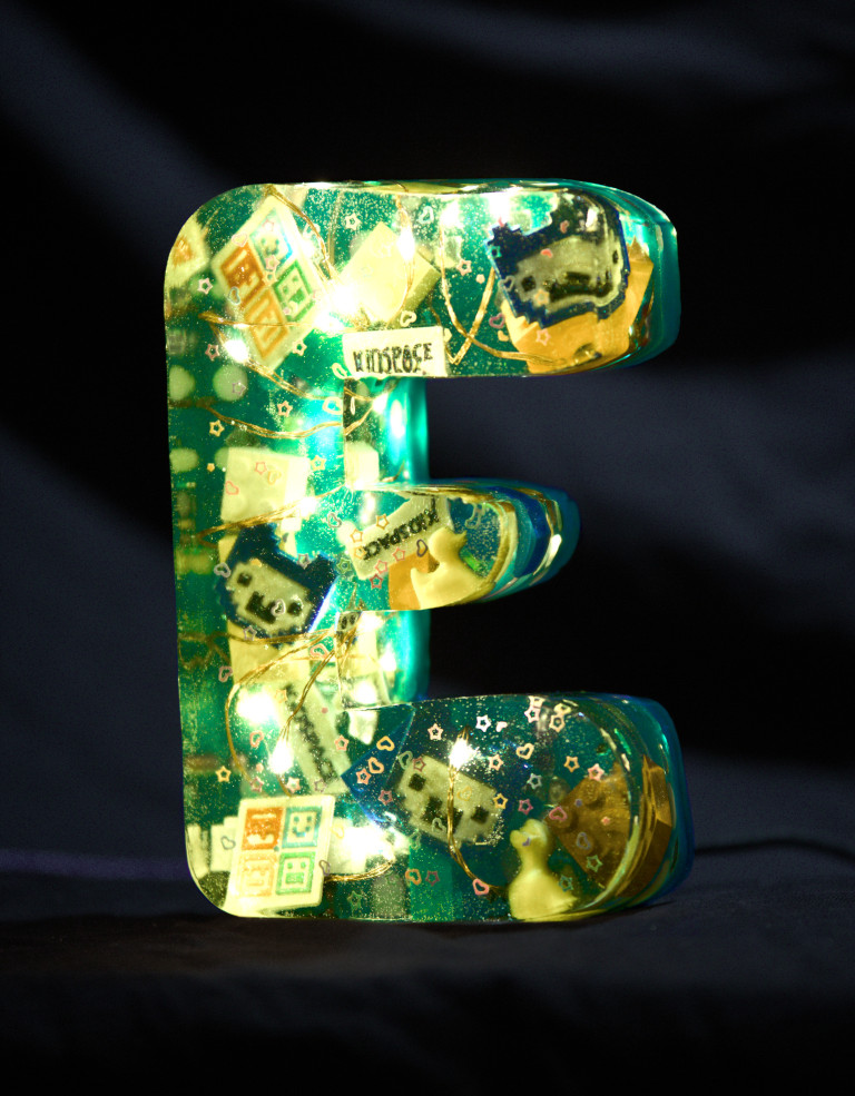 An E cast from epoxy resin. Inside, some fairy lights glow, and colourful glitter stones, Lego bricks, the Jugend Hackt Alpaka logo, the Kidspace logo, little duckies, stars and hearts float around. The letter is turquoise in colour.
        Photo (c) c3awareness | TRim