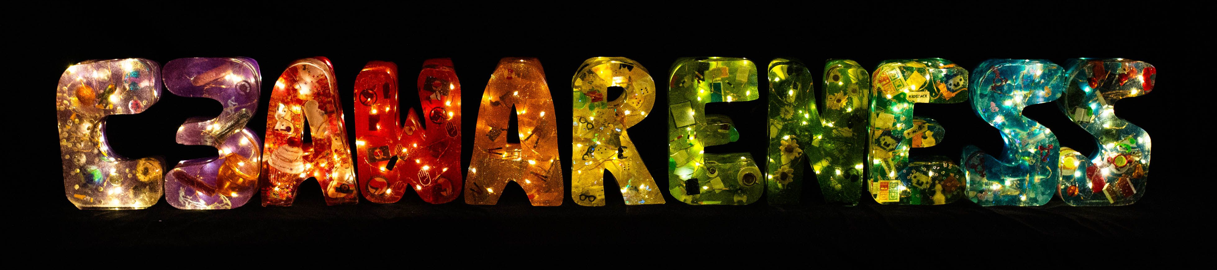 11 letters made of epoxy resin in a rainbow colour gradient. aus Epoxidharz gegossene Buchstaben in einem Regenbogenfarbverlauf. The letters are lit up from the inside with small LEDs and spell C3AWARENESS. Photo (c) c3awareness | TRim